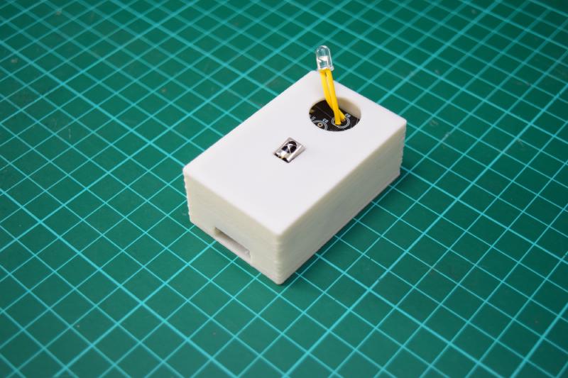 White 3D printed box containing the previous board. On the top side, there is a circular hole about 2 centimetres in diameter. An LED with yellow insulated legs pokes up through it. Below that hole is a small rectangular one, with the infrared sensor poking through it. On the front side of the box, near its floor, is a rectangular hole for a USB cable.