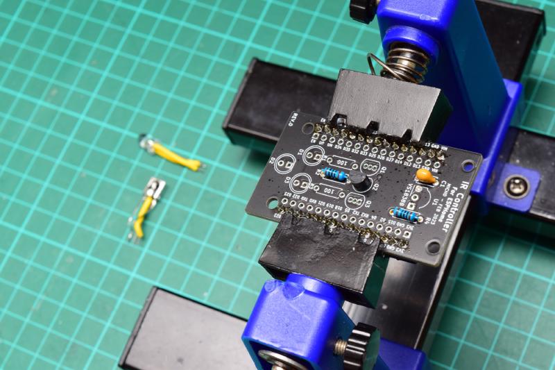IR Controller for ESPHome circuit board in PCB vise. The infrared LED with the long insulated leads, and the infrared receiver with crossed over legs have been removed from the board and lay discarded in the background.