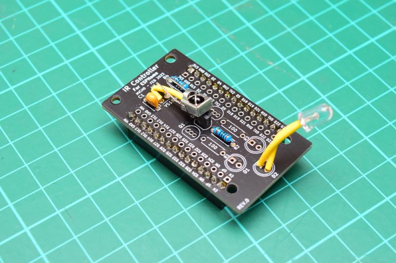 A black circuit board, about 3 by 5.5 centimetres. It is populated with an infrarer receiver which has two pins crossing over, one insulated; a couple of resistors, a TO-92 package MOSFET, a ceramic capacitor, and a clear LED with long insulated leads. On the bottom of the board are pin sockets. The text on the top of the board reads: IR Controller For ESPHome, MICHD - FEB 2023. In the bottom left corner: REV.0