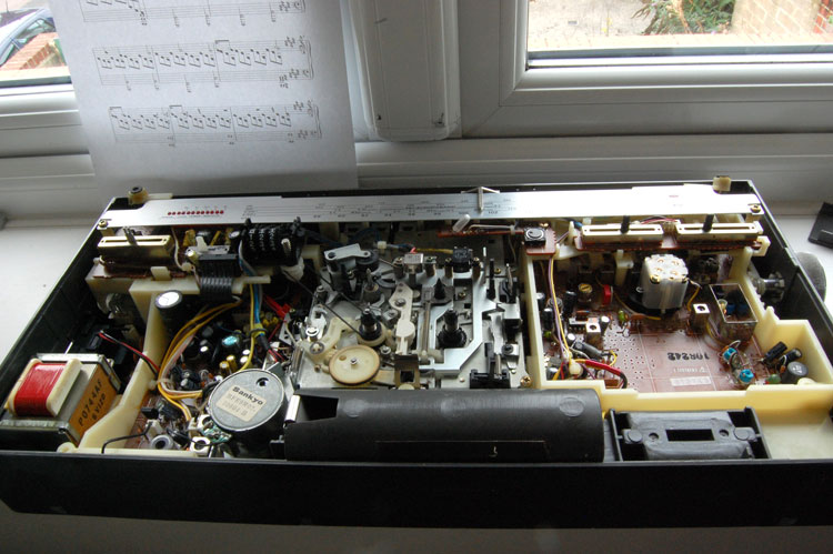 Photo of the radio laying down on its back, the front part of the case taken off. Revealed is the mechanism of the cassette player, various sliders, PCBs with various components on the left and right, and the transformer on the far left.