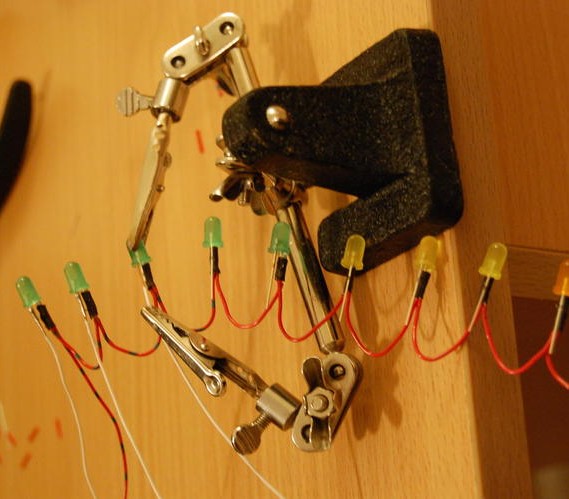 A Helping hand tool holds an LED and a wire, connected to a string of other LEDs by a chain or red wire